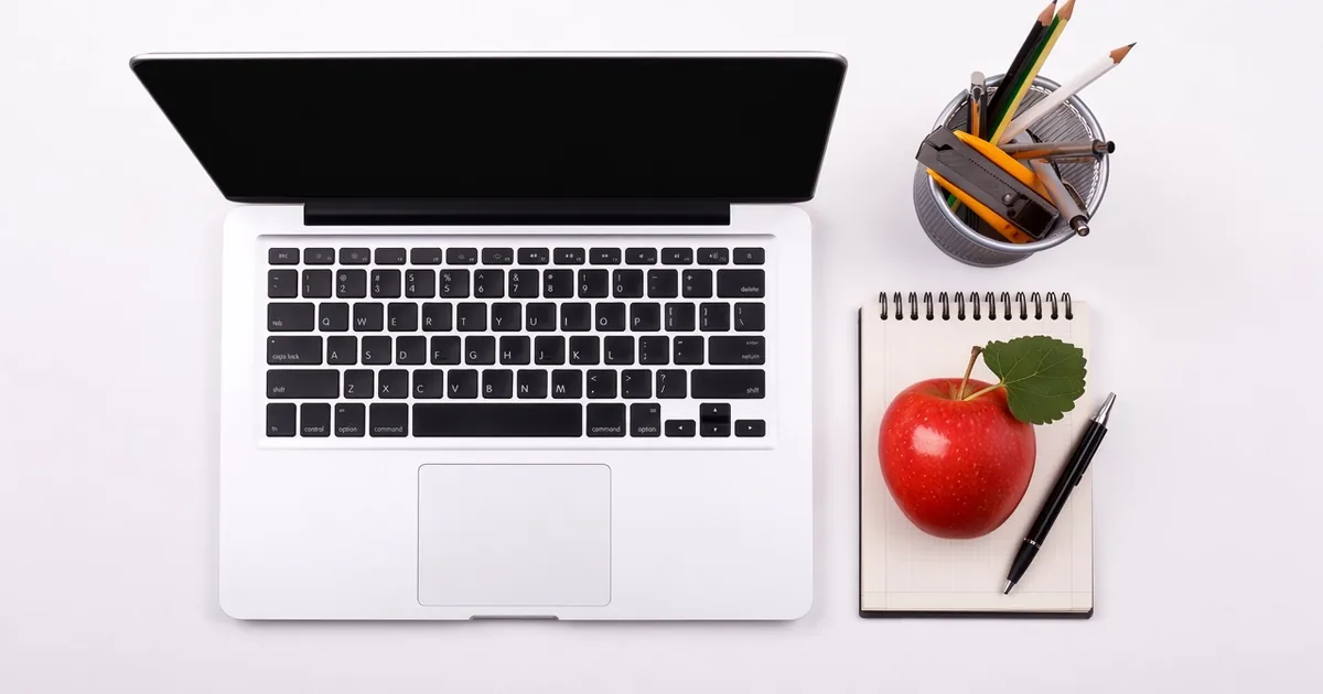 Top-down view of a silver laptop on a white desk beside a spiral notepad with a red apple, a black pen and a cup of pencils.